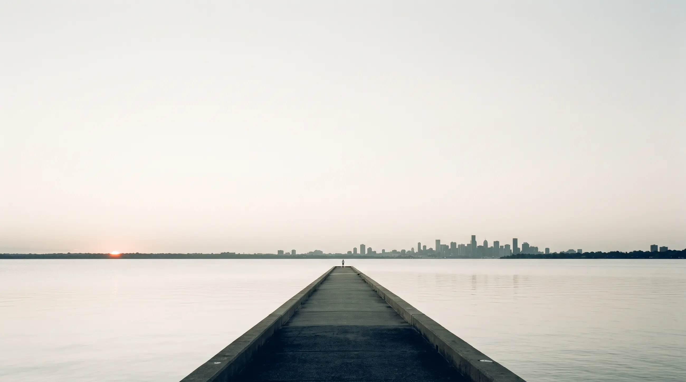 A concrete pier running into still water at first light, a distant skyline low on the horizon