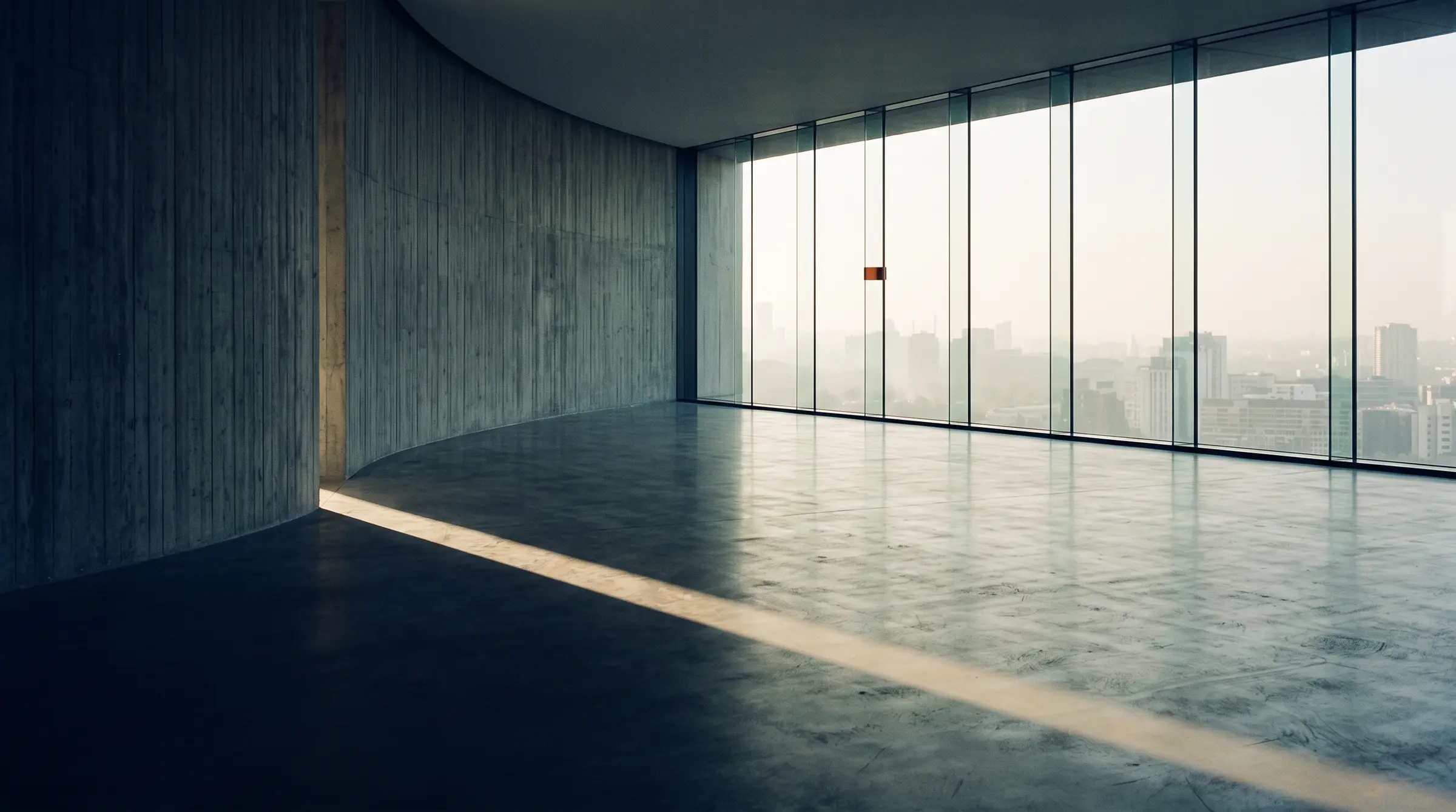 An empty curved concrete hall meeting a full-height window, low sun crossing the floor