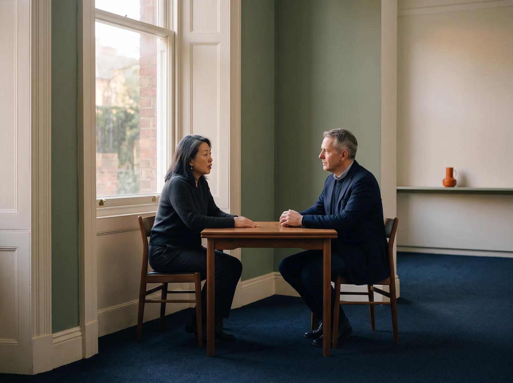 Two senior executives in a one-to-one conversation across a small table beside a tall window